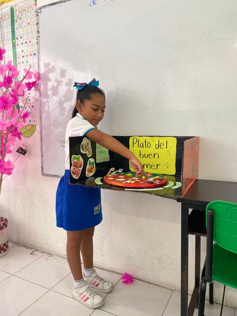 Student in blue skirt holding a handmade cardboard food stand labeled Plato del buen comer in a classroom