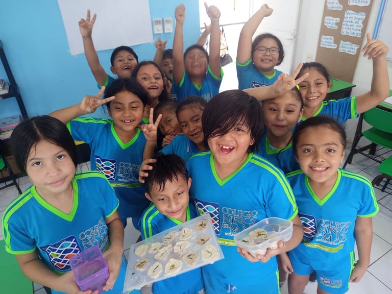 Group of children in matching blue and green uniforms celebrating with raised arms in a classroom, holding painted artworks
