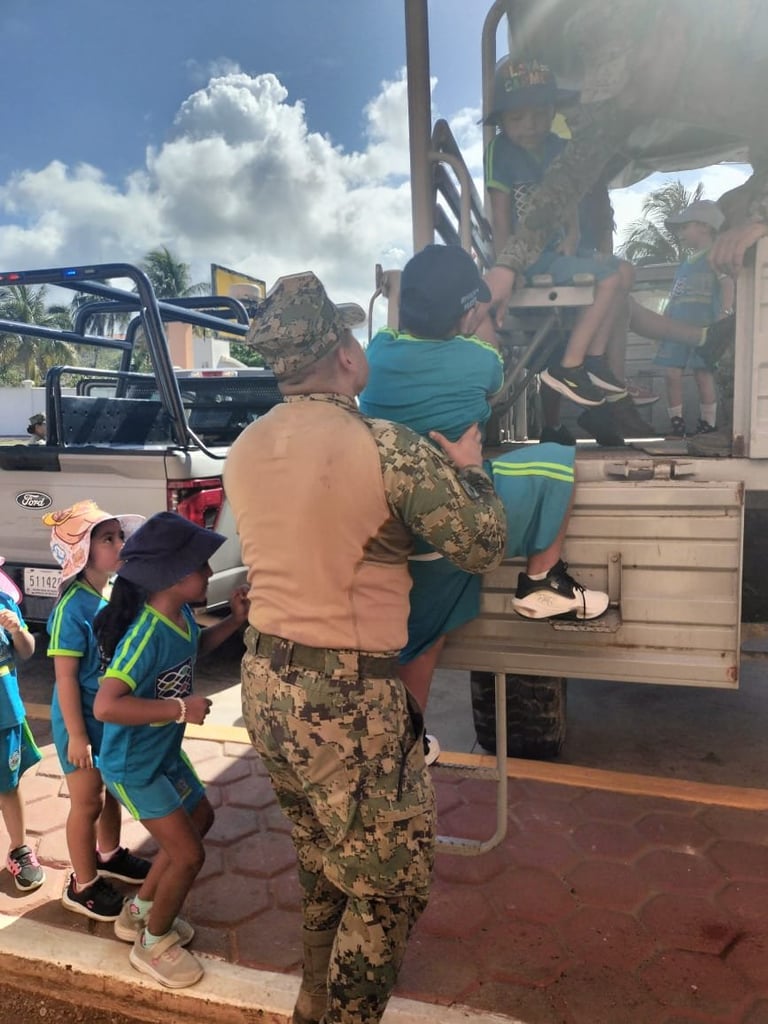 Military personnel in camouflage uniform helping children board a large truck under blue sky with clouds